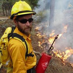 Image of firefighter, Bobby Woelz, lighting a prescribed fire with a drip torch in his hand. 